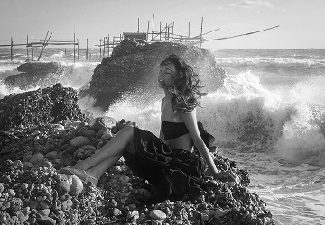 Fekete-fehér fotó egy nőről nyitott térben, aki a hullámzó tenger mellett ül egy sziklán