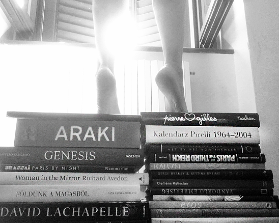 Fekete-fehér fotó egy könyvkupacról, amelyen egy ember mezítlábas lábai állnak egy belső térben