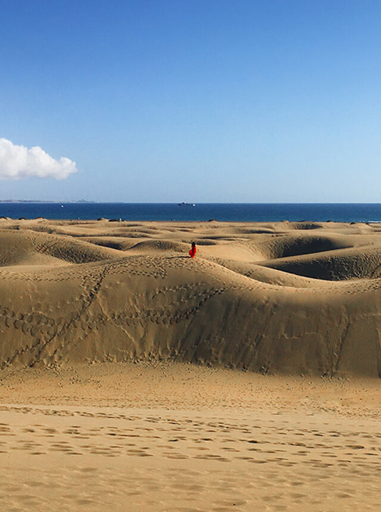 Sivatagi táj homokdűnékkel, a kép közepén egy távoli, piros ruhát viselő ember alakja látható.